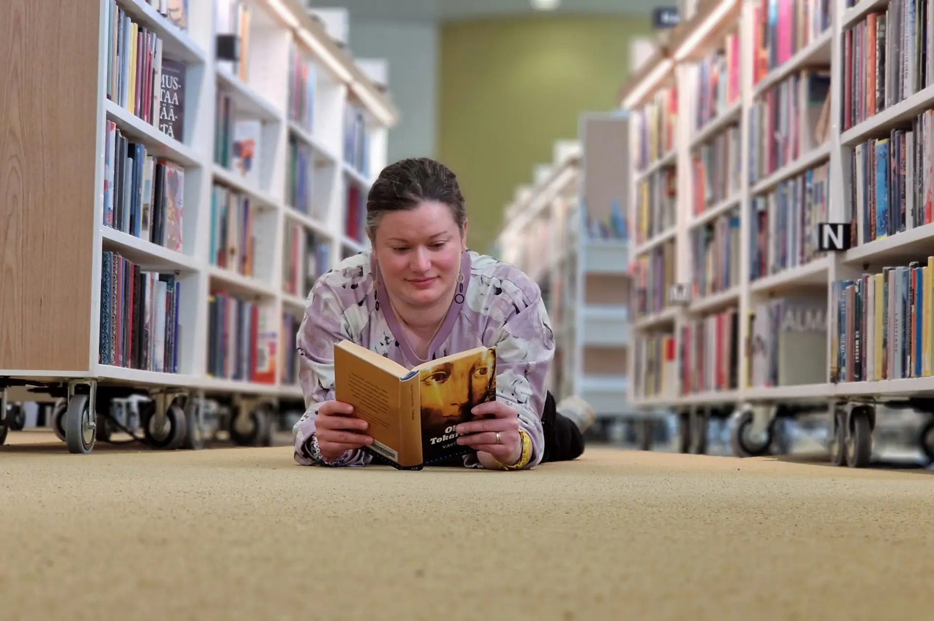 Kuvassa on henkilö lukemassa kirjaston lattialla. Bilden visar en person, som ligger på golvet på bibiliotek och läser. The picture shows a person lying on the floor in a library and reading.