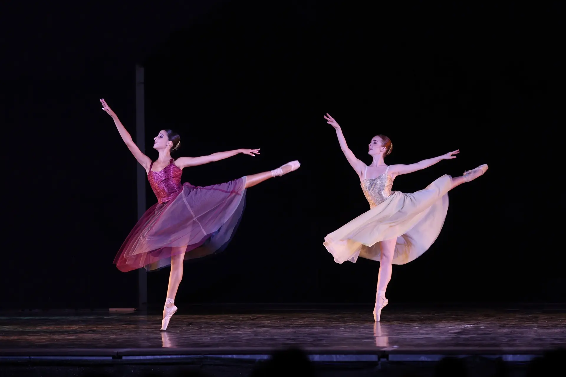 Two ballet dancers performing on stage in flowing dresses, balanced en pointe with extended arms and legs under dramatic theatrical lighting.