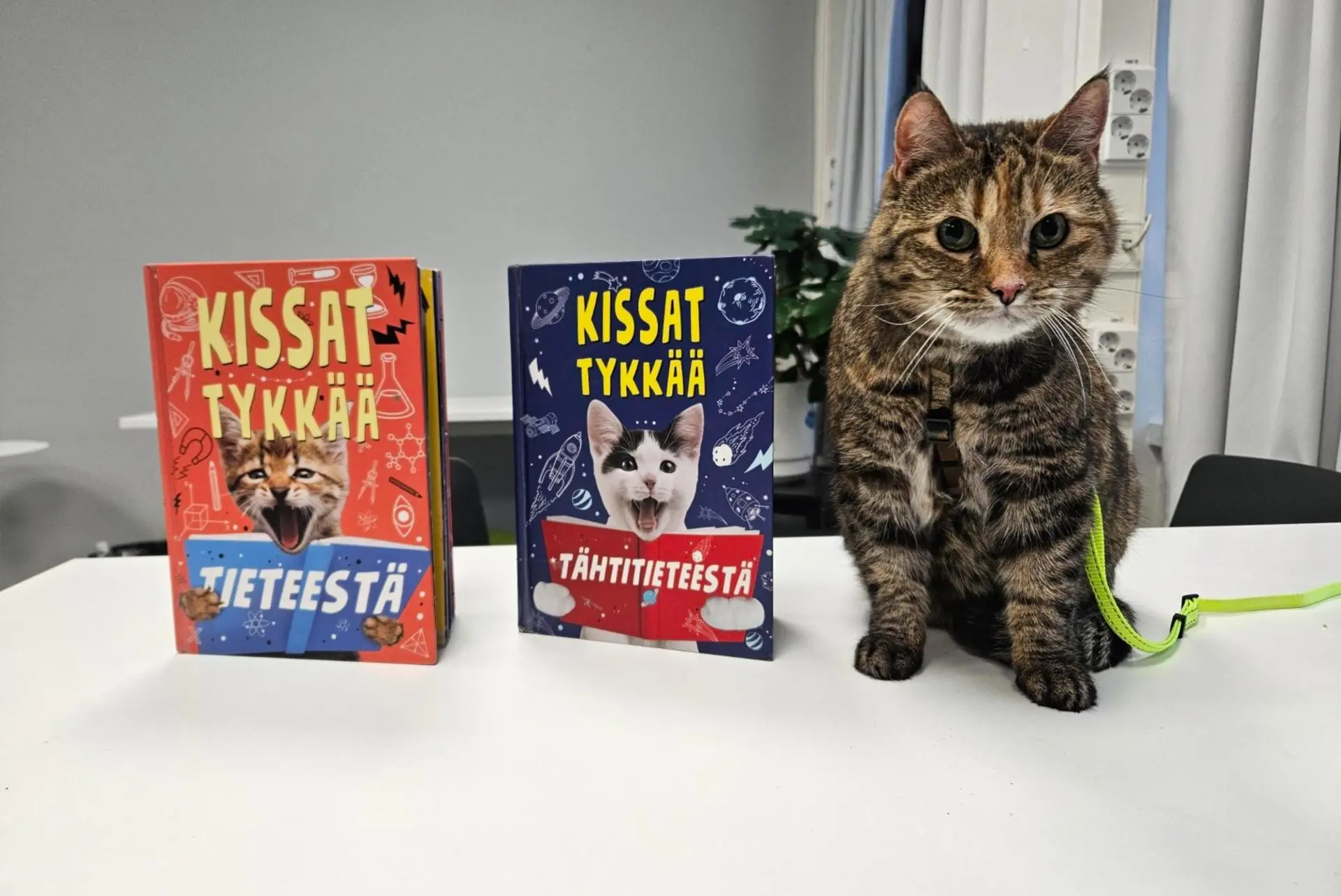 Valkoisella pöydällä istuu raidallinen lukukissa Tuska. Pöydällä Tuskan vieressä on kaksi kissa-aiheista tietokirjaa.