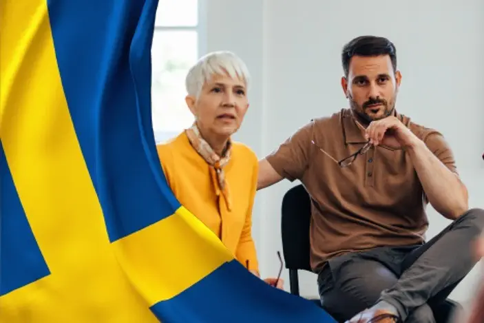 kaksi henkilöä istumassa, kuvan reunassa ruotsin lippu