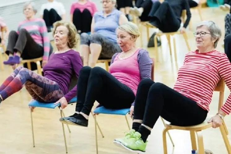 Kolme tuolijumppaajaa tekemässä vatsalihasliikkeitä nostamalla jalkoja koukussa ylös ja pitäen käsillä tukea tuolin takaosastatukea