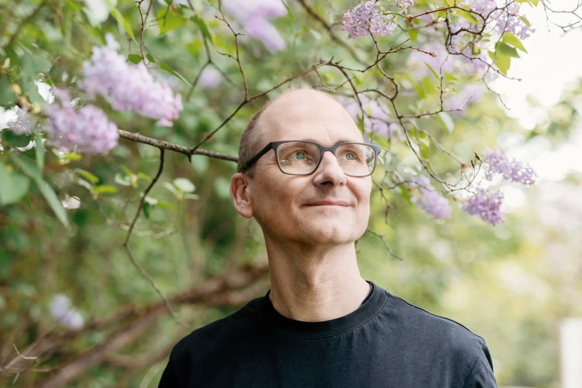 Kirjailija Leo Stranius katsoo yläviistoon ohi kameran ja hymyilee. Hänellä on musta paita ja silmälasit. Stranius seisoo kukkivan puun edessä.