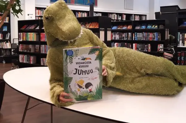 Satukrokotiilipukuun pukeutunut henkilö makaa pöydällä ja pitelee kirjaa