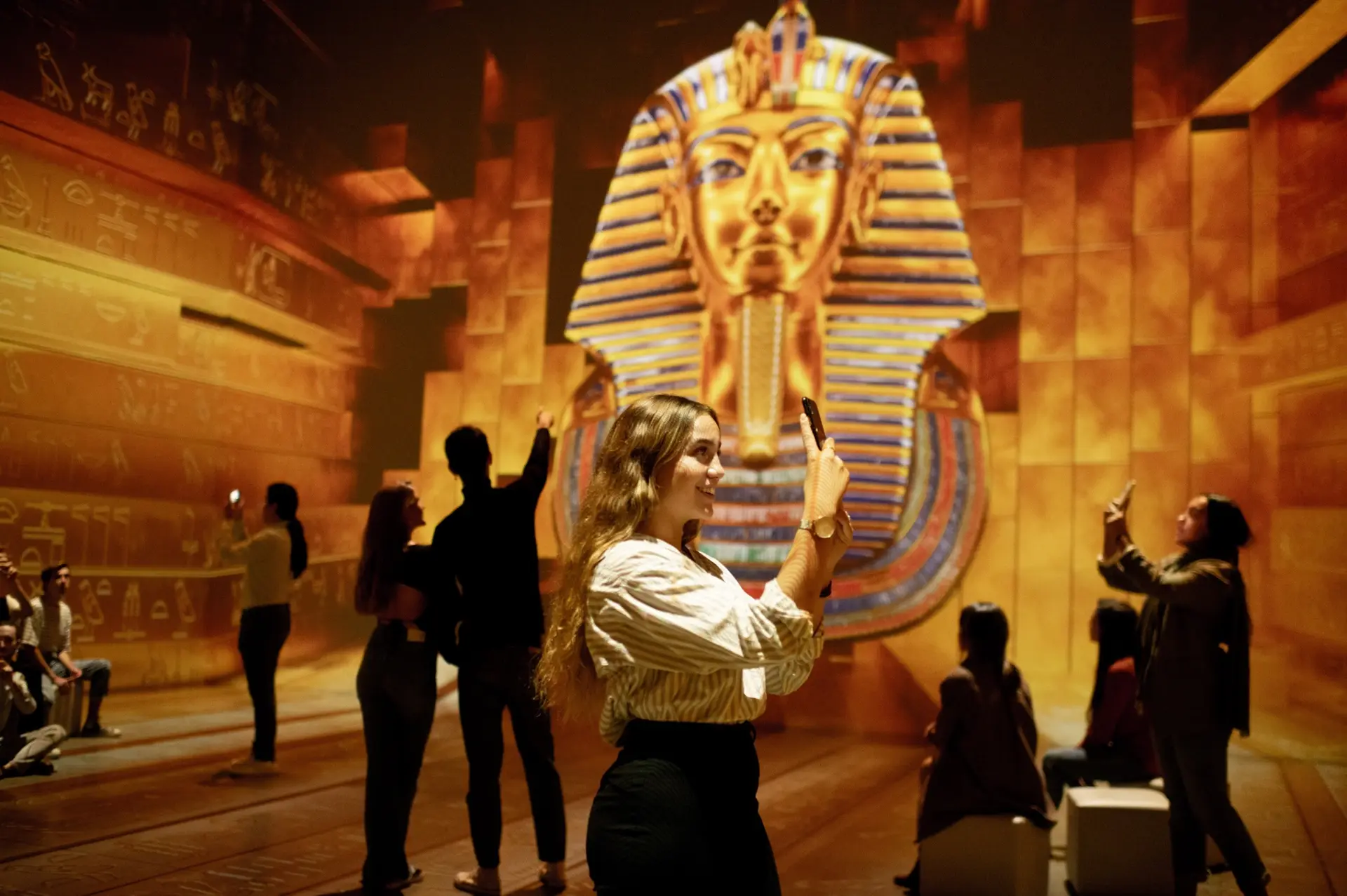 Kävijöitä Tutankhamun – The Immersive Exhibitionin immersiivisessä näyttelytilassa, jossa Tutankhamonin kultainen naamio heijastuu suurena seinälle.