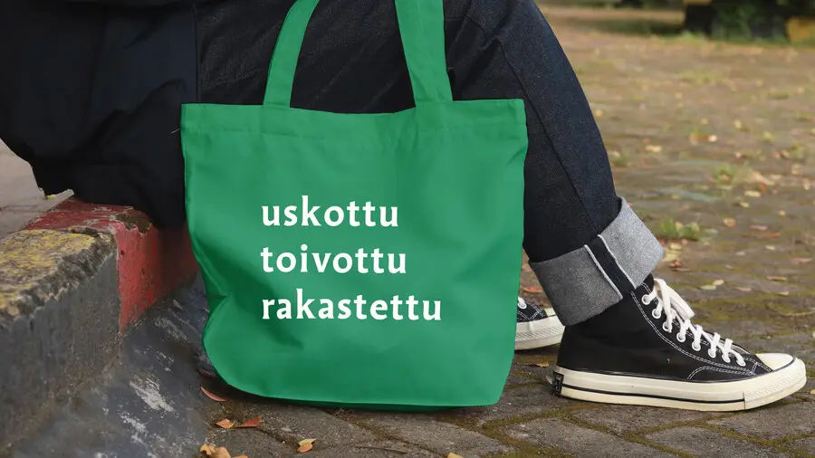 istuvan henkilön jalat ja kangaskassi, jossa teksti uskottu, toivottu, rakastettu