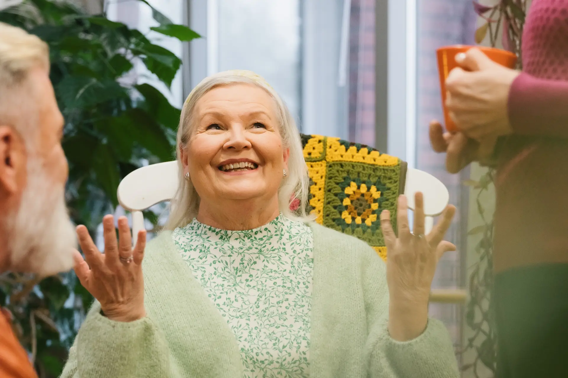 Kuvassa keskellä hymyilevä seniori, jonka vieressä kaksi muuta henkilöä, joista toisella kahvikuppi kädessä